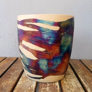 Puede incluir: Un macetero de cerámica con base blanca y glaseado iridiscente azul, morado y cobre. El glaseado tiene un efecto craquelado, lo que le da al macetero un aspecto único y rústico.