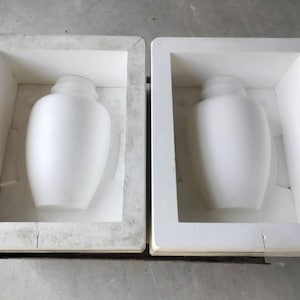 Könnte beinhalten: Zwei weiße, vasenförmige Objekte werden in rechteckigen Formen gezeigt. Die Vasen haben einen abgerundeten Körper und einen schmalen Hals. Die Formen sind weiß und haben ein einfaches, kastenförmiges Design. Der Hintergrund ist eine hellgraue Oberfläche.