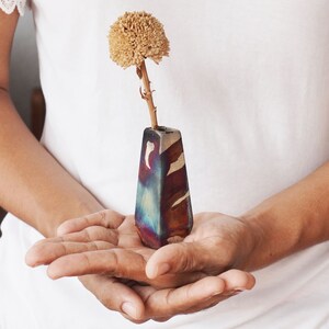 Puede incluir: Un pequeño jarrón de cerámica hecho a mano con un esmalte azul, morado y marrón. El jarrón contiene una sola flor seca con un tallo marrón.