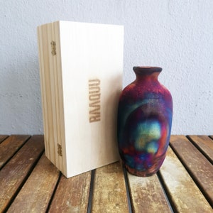 May include: A small, colorful ceramic vase with a swirling pattern of red, blue, and gold. The vase is sitting on a wooden surface next to a wooden box with the word "RMQQU" engraved on it.