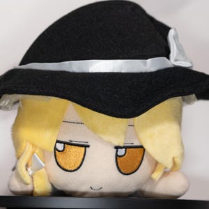 May include: A plush doll featuring a character with yellow hair and orange eyes, wearing a black hat with a white ribbon. The doll has a neutral expression and is made of soft fabric, likely for collectors or fans.