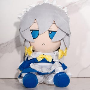 May include: A plush doll dressed as a maid. The doll features grey hair, blue eyes, and a blue and white dress with yellow bows. The doll is seated and has a neutral expression.