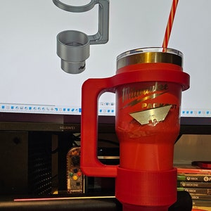 Puede incluir: Un vaso t&eacute;rmico rojo con asa y pajita a rayas rojas y blancas. El vaso tiene el logotipo de Milwaukee Packout. Tambi&eacute;n se ve un portavasos impreso en 3D. El texto "Rigg3d" est&aacute; impreso en la parte inferior del vaso.