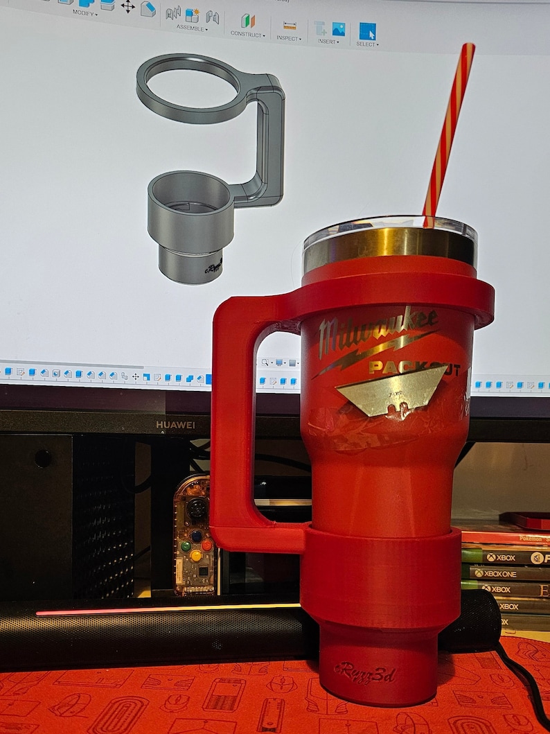 Puede incluir: Un vaso t&eacute;rmico rojo con asa y pajita a rayas rojas y blancas. El vaso tiene el logotipo de Milwaukee Packout. Tambi&eacute;n se ve un portavasos impreso en 3D. El texto "Rigg3d" est&aacute; impreso en la parte inferior del vaso.