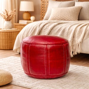 Puede incluir: Un otomán redondo de cuero rojo con costuras blancas. El otomán cilíndrico está sobre una alfombra estampada. El fondo incluye una cama con cabecero tejido y una mesita de noche con una lámpara.