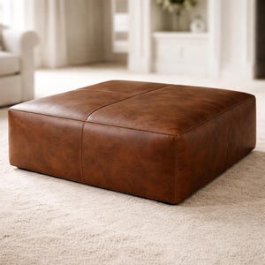 May include: A square, brown leather ottoman with a stitched design. The ottoman is a warm, medium brown color and sits on a light-colored carpet. The design features four distinct sections, adding to its visual appeal. This piece is suitable for living room furniture.