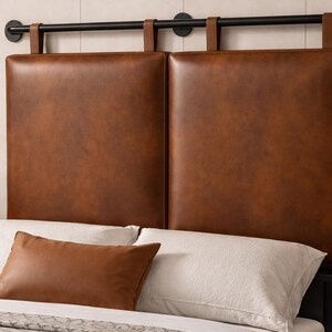 May include: A brown leather headboard with a black metal frame. The headboard features two large, square panels. A matching brown leather pillow rests on the bed, which has white sheets and a patterned blanket.