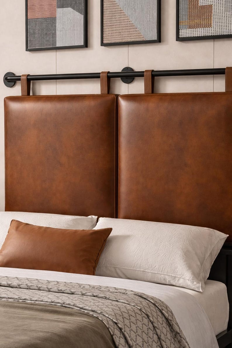 May include: A brown leather headboard with a black metal frame. The headboard features two large, square panels. A matching brown leather pillow rests on the bed, which has white sheets and a patterned blanket.