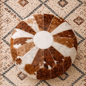 Puede incluir: Puf redondo de patchwork de piel de vaca en tonos marrones y blancos. El puf tiene un centro circular blanco y está rodeado de piezas triangulares de piel. El diseño recuerda a un balón de fútbol. Está colocado sobre una alfombra estampada.