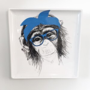 May include: A white, rectangular ceramic tray featuring a whimsical illustration of a monkey wearing blue glasses and a blue headband. The monkey is drawn in black and white, with the blue accents providing a pop of color.