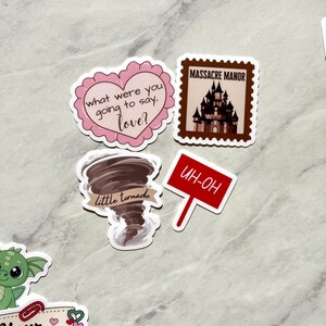Puede incluir: Una colecci&oacute;n de pegatinas con varios dise&ntilde;os. Una pegatina es un coraz&oacute;n rosa con el texto "what were you going to say, love?". Otra es un sello marr&oacute;n con un castillo y las palabras "MASSACRE MANOR". Otras pegatinas incluyen un tornado y un letrero rojo que dice "UH-OH".