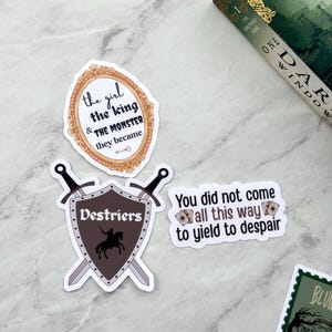 Op de afbeelding: Een verzameling stickers met citaten en afbeeldingen. E&eacute;n sticker toont de tekst "the girl & the king & THE MONSTER they became" in een sierlijke lijst. Een andere toont een schild met een ridder en het woord "Destriers".