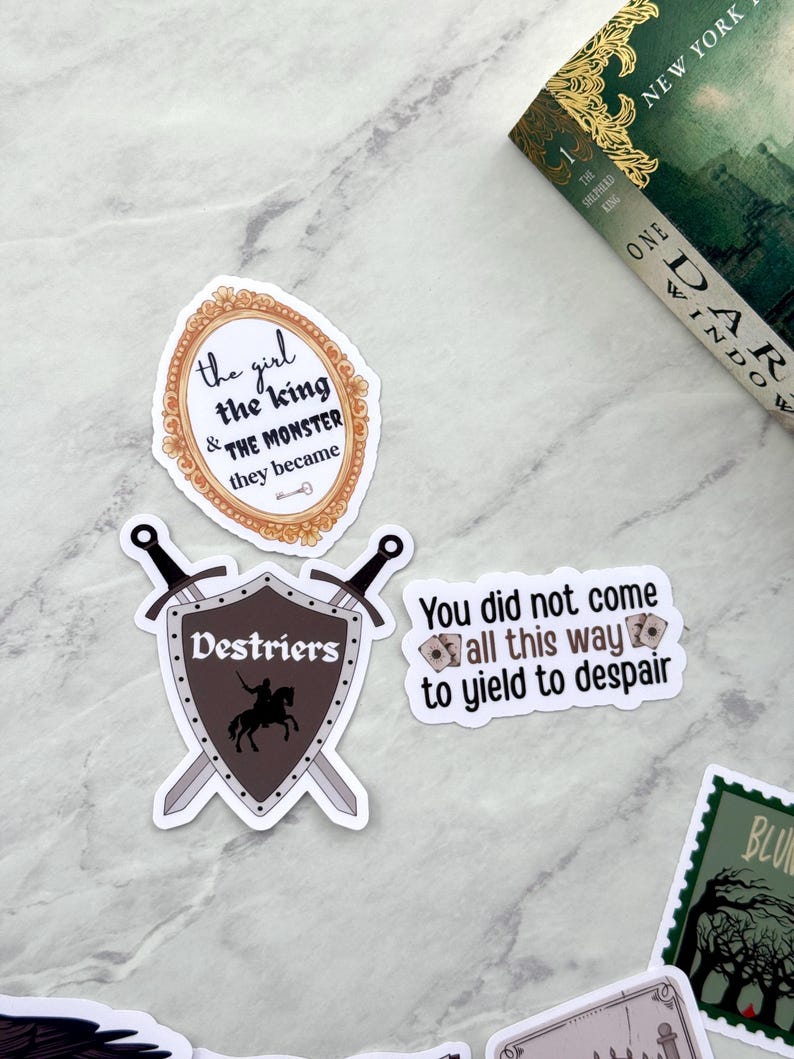 Op de afbeelding: Een verzameling stickers met citaten en afbeeldingen. E&eacute;n sticker toont de tekst "the girl & the king & THE MONSTER they became" in een sierlijke lijst. Een andere toont een schild met een ridder en het woord "Destriers".