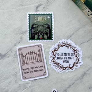 Op de afbeelding: Een verzameling stickers met dark fantasy thema's. E&eacute;n sticker toont het woord "BLUNDER" boven een bossc&egrave;ne. Een andere luidt "HE CAME FOR THE GIRL AND GOT THE MONSTER INSTEAD." Een derde zegt "having hope does not make me delusional."