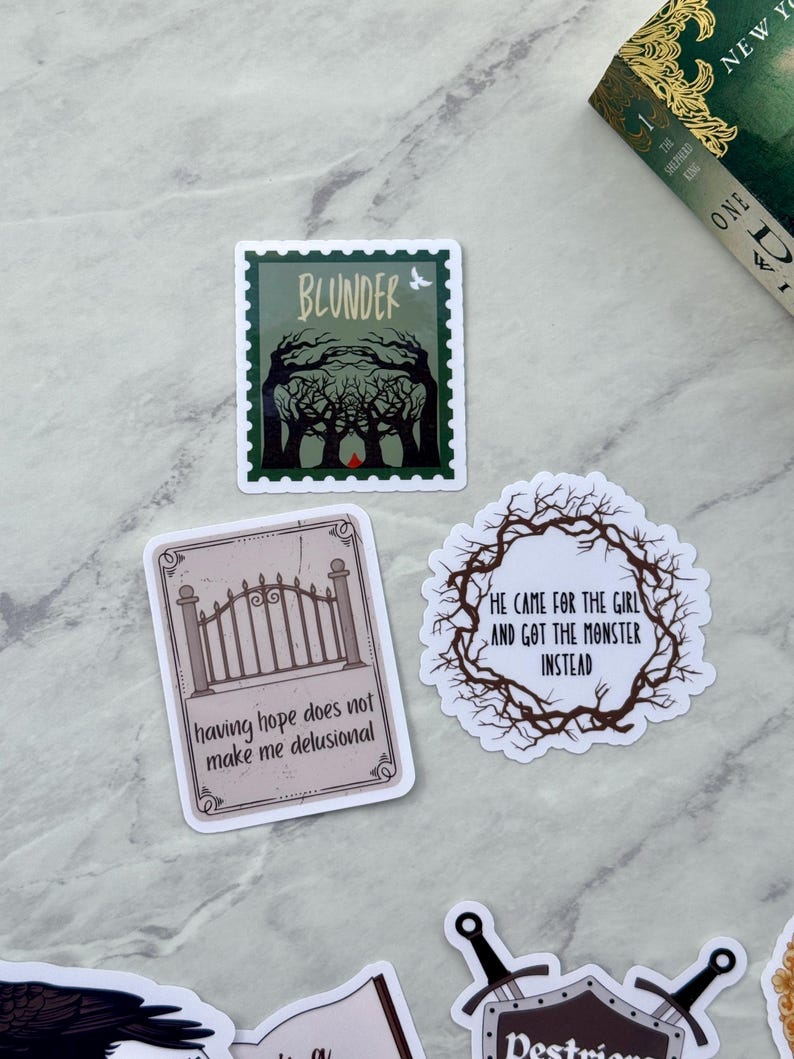 Op de afbeelding: Een verzameling stickers met dark fantasy thema's. E&eacute;n sticker toont het woord "BLUNDER" boven een bossc&egrave;ne. Een andere luidt "HE CAME FOR THE GIRL AND GOT THE MONSTER INSTEAD." Een derde zegt "having hope does not make me delusional."
