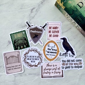 Op de afbeelding: Een verzameling stickers met fantasiethema's met verschillende ontwerpen en citaten. Stickers zijn onder meer een raaf, een schild met gekruiste zwaarden en zinnen als "Be Wary Be Clever Be Good". Er is ook een boek met een groene kaft aanwezig.