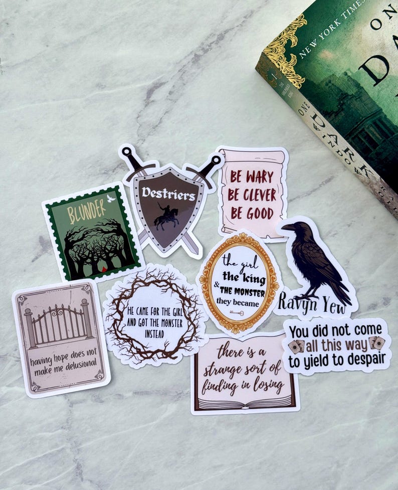 Op de afbeelding: Een verzameling stickers met fantasiethema's met verschillende ontwerpen en citaten. Stickers zijn onder meer een raaf, een schild met gekruiste zwaarden en zinnen als "Be Wary Be Clever Be Good". Er is ook een boek met een groene kaft aanwezig.