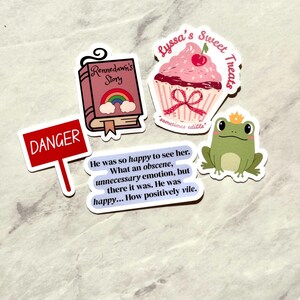 Puede incluir: Una colecci&oacute;n de pegatinas con varios dise&ntilde;os. Una pegatina presenta una magdalena rosa con una cereza y el texto "Lyssa's Sweet Treats". Otra muestra un libro con un arco&iacute;ris y el texto "Rennedawn's Story". Otras pegatinas incluyen una rana, un letrero de "Danger" y una cita.