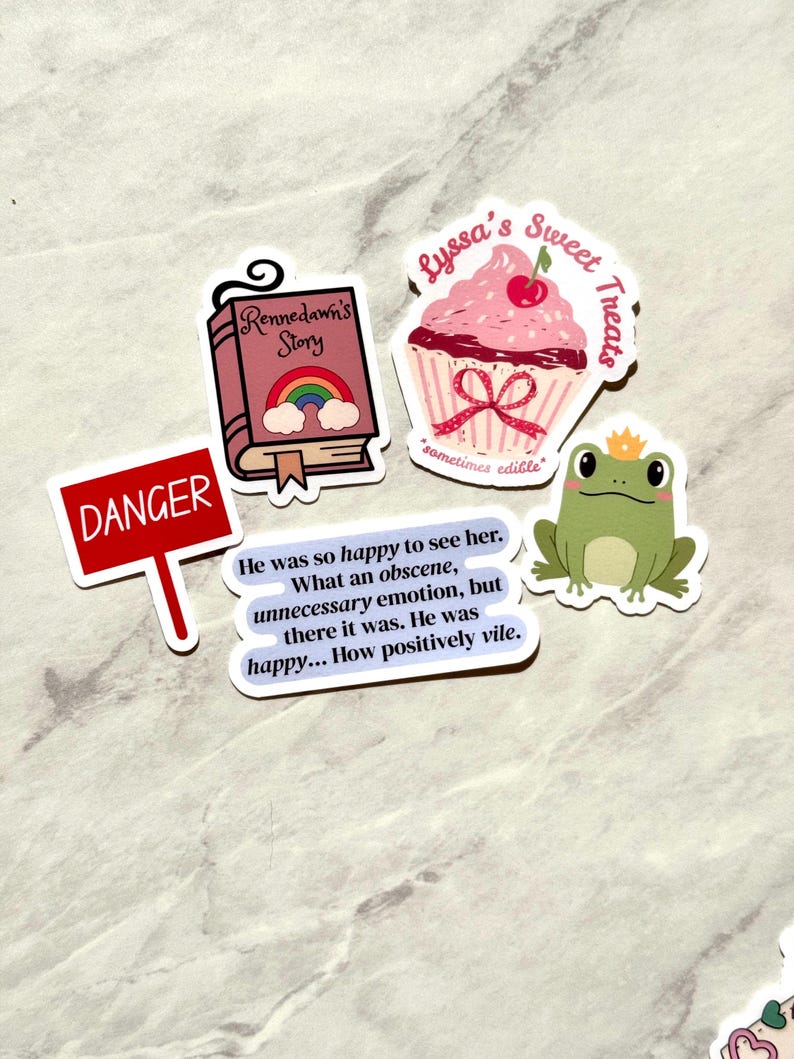 Puede incluir: Una colecci&oacute;n de pegatinas con varios dise&ntilde;os. Una pegatina presenta una magdalena rosa con una cereza y el texto "Lyssa's Sweet Treats". Otra muestra un libro con un arco&iacute;ris y el texto "Rennedawn's Story". Otras pegatinas incluyen una rana, un letrero de "Danger" y una cita.