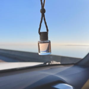 May include: A clear glass bottle with a brown cap hanging from a string in a car. The bottle is filled with a light pink liquid. The bottle is hanging from the rearview mirror.