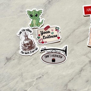 Puede incluir: Una colecci&oacute;n de pegatinas caprichosas que incluyen un drag&oacute;n verde, una calavera con una vela y un letrero que dice "Edwin's Cauldron Brew". Otras pegatinas incluyen las frases "I am the villain, the dark fears me" y "Your Evilness".