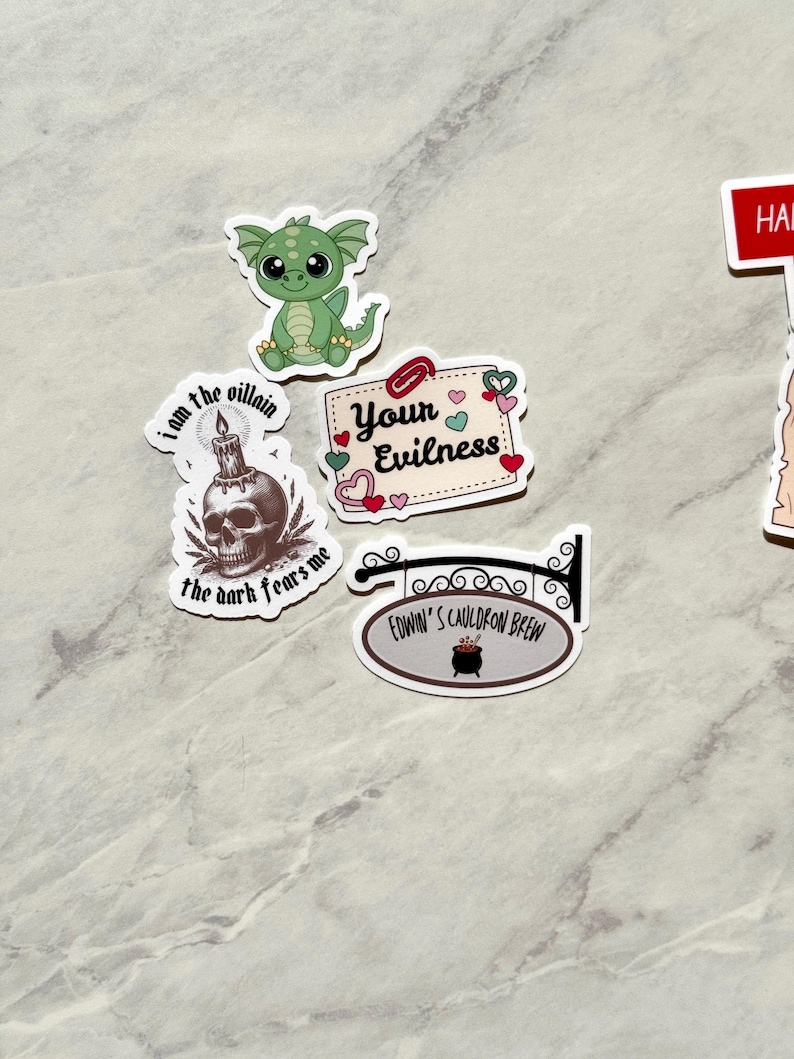 Puede incluir: Una colecci&oacute;n de pegatinas caprichosas que incluyen un drag&oacute;n verde, una calavera con una vela y un letrero que dice "Edwin's Cauldron Brew". Otras pegatinas incluyen las frases "I am the villain, the dark fears me" y "Your Evilness".