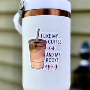 May include: White tumbler with a sticker that says "I like my coffee icy and my books spicy". The sticker features a cartoon drawing of an iced coffee.