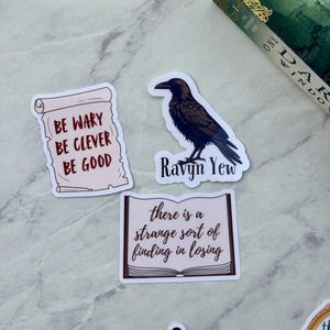 Op de afbeelding: Een verzameling stickers met citaten en illustraties. E&eacute;n sticker toont een zwarte raaf met de tekst "Ravyn Yew". Een andere luidt "Be wary, be clever, be good". Een derde zegt "there is a strange sort of finding in losing."