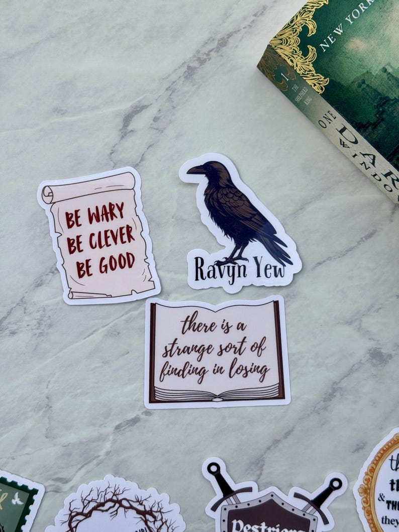 Op de afbeelding: Een verzameling stickers met citaten en illustraties. E&eacute;n sticker toont een zwarte raaf met de tekst "Ravyn Yew". Een andere luidt "Be wary, be clever, be good". Een derde zegt "there is a strange sort of finding in losing."