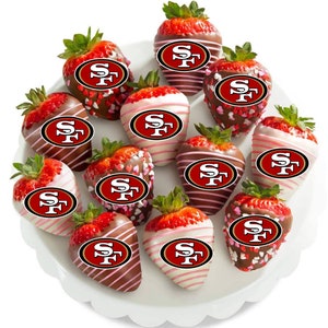以下が含まれることがあります： 白いスカラップエッジの皿には、チョコレートでコーティングされたイチゴが載っています。 イチゴは、白、ミルク、ダークチョコレートに浸され、サンフランシスコ49ersのロゴで飾られ、一部はハート型のスプリンクルが振りかけられています。
