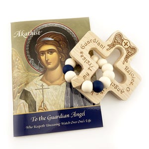 May include: A wooden cross-shaped baby teether with engraved text and angel wings, paired with a book titled "Akathist To the Guardian Angel." The teether features white and navy blue beads. The book has an illustration of an angel.