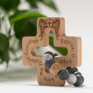 May include: Wooden cross with a silicone beaded bracelet attached. The cross has the words "Rejoice, O Angel" and "My Lord, Thy Precious Guardian!" engraved on it. The cross has a carved angel wing design with a cross in the center.