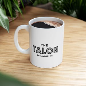 Peut inclure: Une tasse à café en céramique blanche avec du texte noir qui dit "THE TALON SMALLVILLE, KS". La tasse est remplie de café et est posée sur une table en bois.