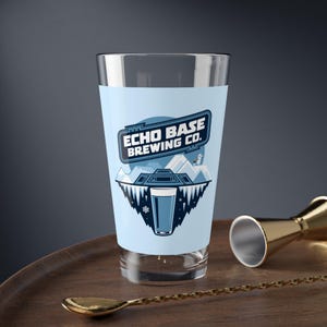 Könnte beinhalten: Ein blaues und weißes Glas-Pint-Glas mit dem Text "Echo Base Brewing Co." und einer Grafik eines Raumschiffs und schneebedeckter Berge.