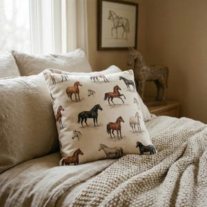 May include: A decorative pillow with a beige background featuring various horse illustrations in brown, black, and white. The pillow is placed on a bed with beige linens and a knitted throw. A framed horse sketch hangs on the wall.