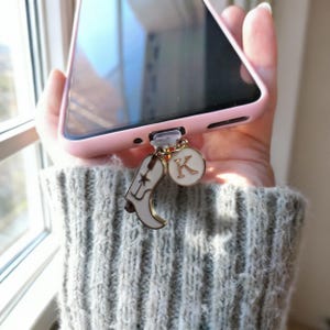 May include: A pink phone case with a charm featuring a white cowboy boot and a gold-rimmed circle with the letter "K". The charm is attached to the phone's charging port.