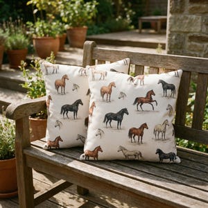 May include: Two square throw pillows with a cream background and a repeating pattern of various horse breeds in brown, black, and tan. The pillows are placed on a wooden bench outdoors.