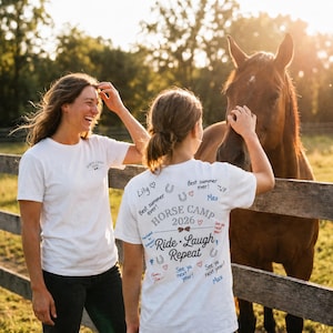 Könnte beinhalten: Zwei Personen tragen weiße T-Shirts mit dem Text "HORSE CAMP 2026" und "Ride, Laugh, Repeat", die neben einem braunen Pferd stehen. Die Shirts haben handschriftliche Notizen und Zeichnungen. Der Hintergrund umfasst einen Holzzaun und ein Feld.