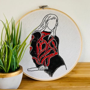 May include: Embroidered hoop art featuring a black and red design of a person holding a microphone. The person has long hair and is wearing a black outfit with red embroidery.