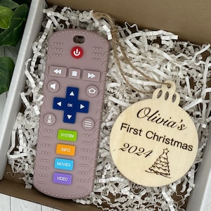 May include: A light brown wooden ornament with the text "Olivia's First Christmas 2024" and a small Christmas tree design. The ornament is in a white box with brown paper shredding. A pink silicone remote control is also in the box.