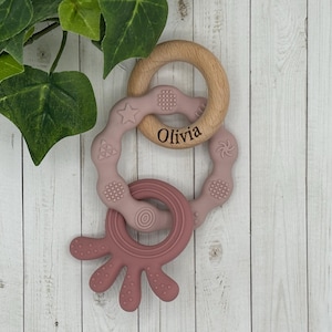 May include: A set of three baby teethers in pink and brown. The teethers are made of silicone and wood. The wooden teether is engraved with the name "Olivia".