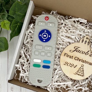 Könnte beinhalten: Eine weiße und graue Fernbedienung mit blauen und grünen Tasten befindet sich in einer Geschenkbox mit weißem Seidenpapier. Ein Holz-Anhänger mit dem Text "Jacob's First Christmas 2024" ist ebenfalls in der Box.