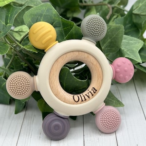 May include: A baby teether with a wooden center and a cream-colored outer ring. The teether has six colorful silicone attachments in yellow, gray, pink, and purple. The name "Olivia" is engraved on the wooden center. The teether is designed for babies.