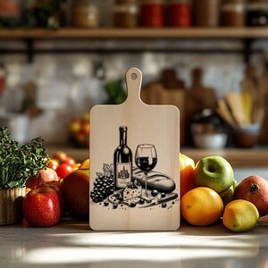 Op de afbeelding: Een houten snijplank met een zwart-witte illustratie van een fles wijn, een glas wijn, druiven, kaas en een stokbrood. De plank heeft een handvat en is gemaakt van natuurlijk hout.