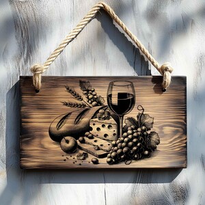 Op de afbeelding: Een houten bord met een gebrande afwerking heeft een handgetekend ontwerp van een glas rode wijn, een brood, kaas en druiven. Het bord heeft een touwbevestiging.