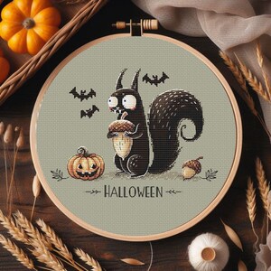 May include: A Halloween-themed cross-stitch design featuring a black squirrel holding an acorn, a jack-o'-lantern, and bats. The text "HALLOWEEN" is stitched below the squirrel. The design is set in a wooden embroidery hoop.
