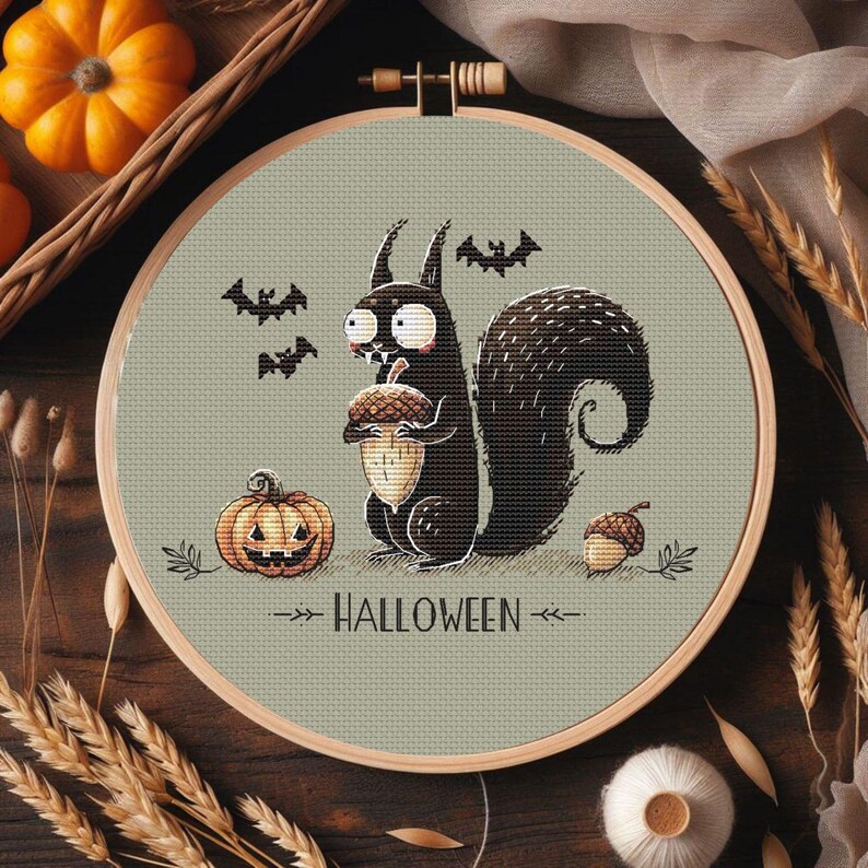 May include: A Halloween-themed cross-stitch design featuring a black squirrel holding an acorn, a jack-o'-lantern, and bats. The text "HALLOWEEN" is stitched below the squirrel. The design is set in a wooden embroidery hoop.