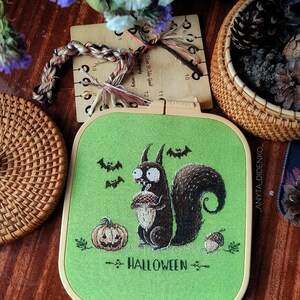 May include: A Halloween-themed cross-stitch embroidery hoop featuring a cartoon squirrel holding an acorn. The design includes a pumpkin, bats, and the word "HALLOWEEN" in green and brown on a green background. The hoop is set against a wooden surface.
