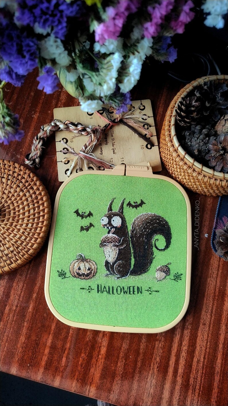 May include: A Halloween-themed cross-stitch embroidery hoop featuring a cartoon squirrel holding an acorn. The design includes a pumpkin, bats, and the word "HALLOWEEN" in green and brown on a green background. The hoop is set against a wooden surface.