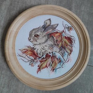Peut inclure: Une broderie au point de croix encadr&eacute;e repr&eacute;sentant un petit lapin blanc assis parmi des feuilles d'automne color&eacute;es. Le lapin a des yeux bruns et une queue touffue.